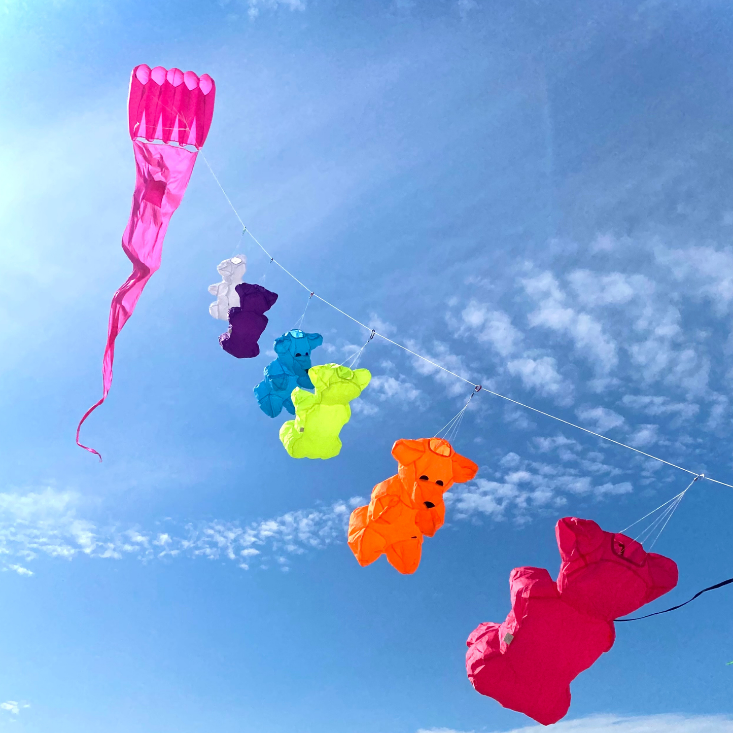 Colorful bear-shaped line laundry flying on a pink lifter kite with a long tail against a blue sky with some clouds.