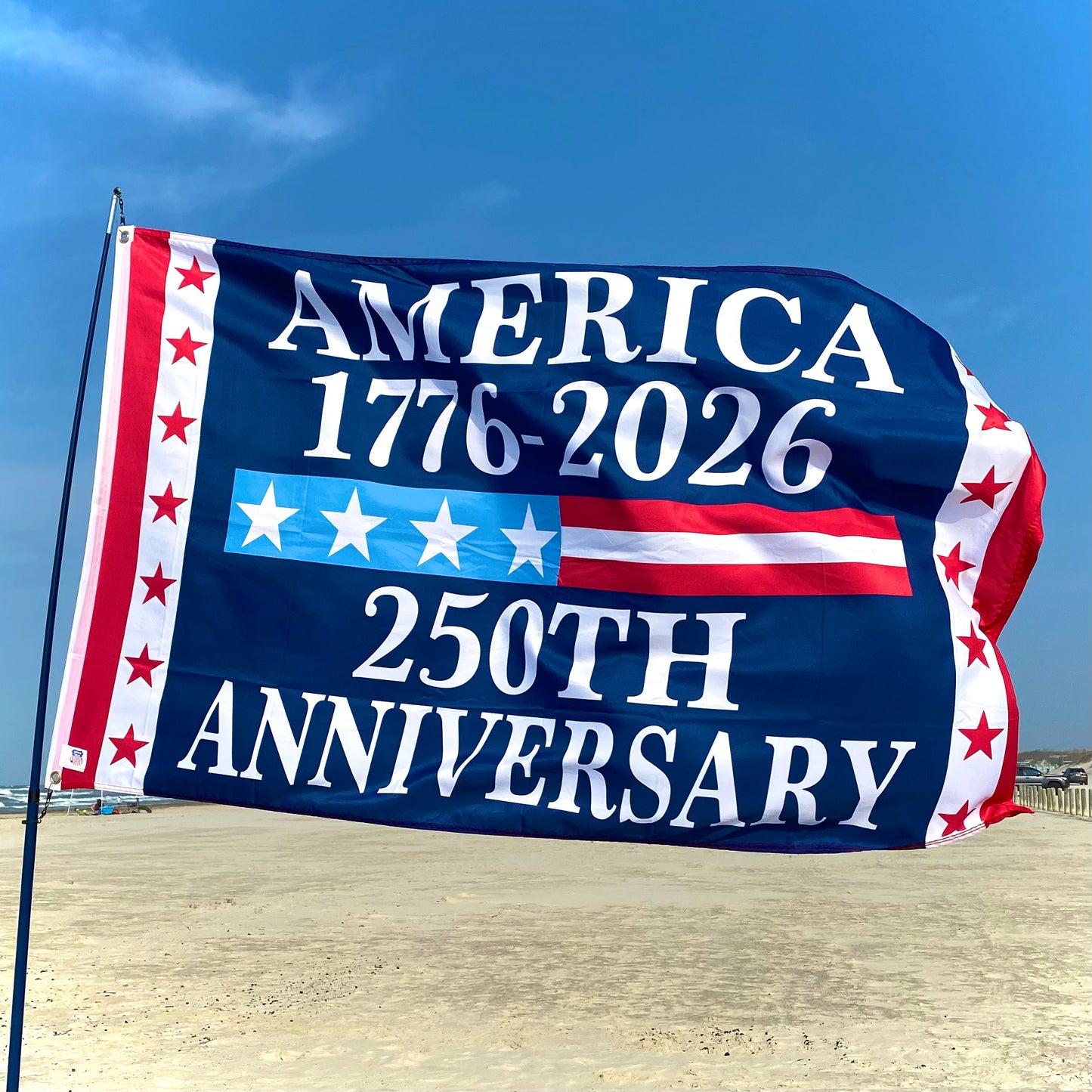 Flag celebrating America's 250th anniversary on a beach with blue sky.