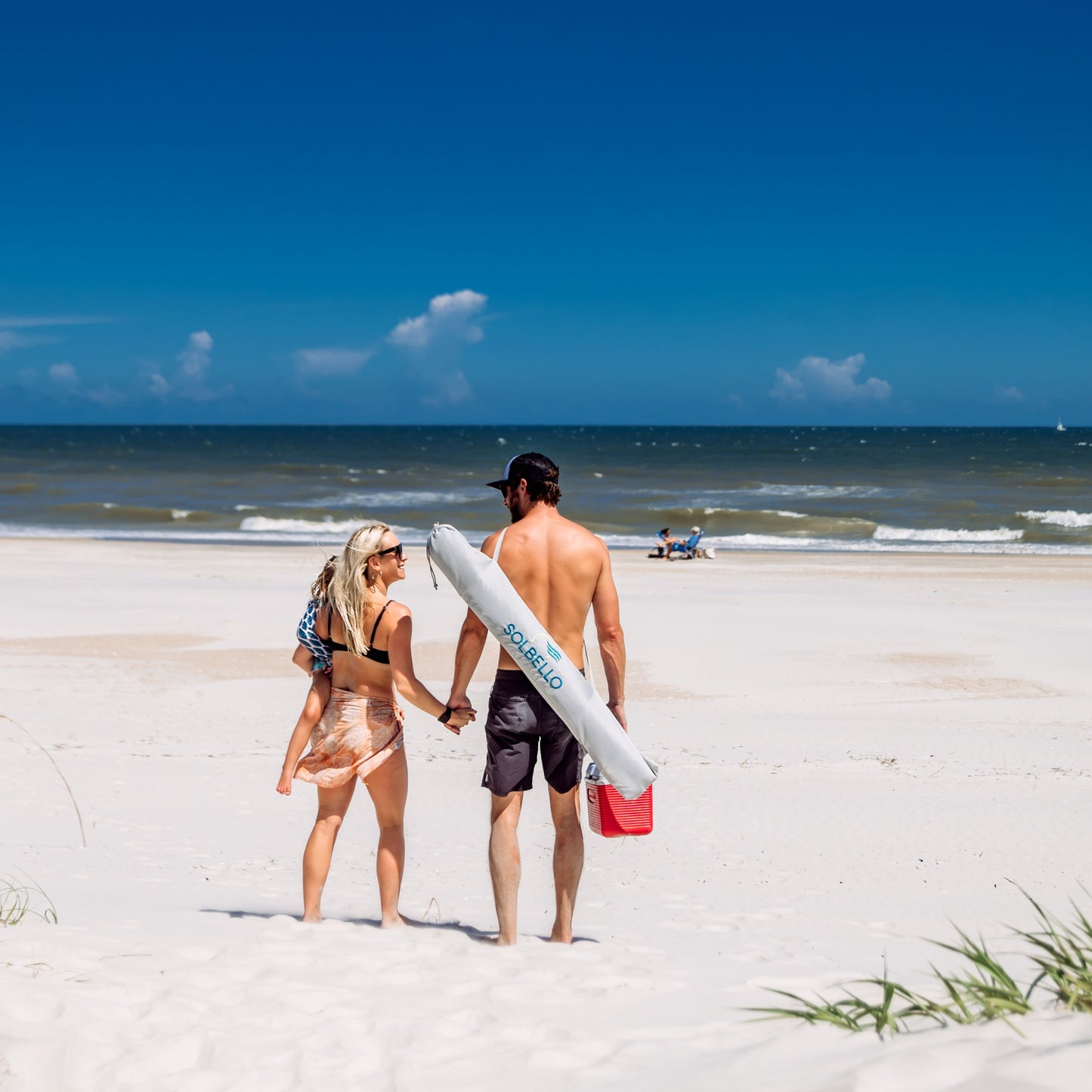 Two people walking on a sandy beach with a clear blue sky carrying a Solbello bag.