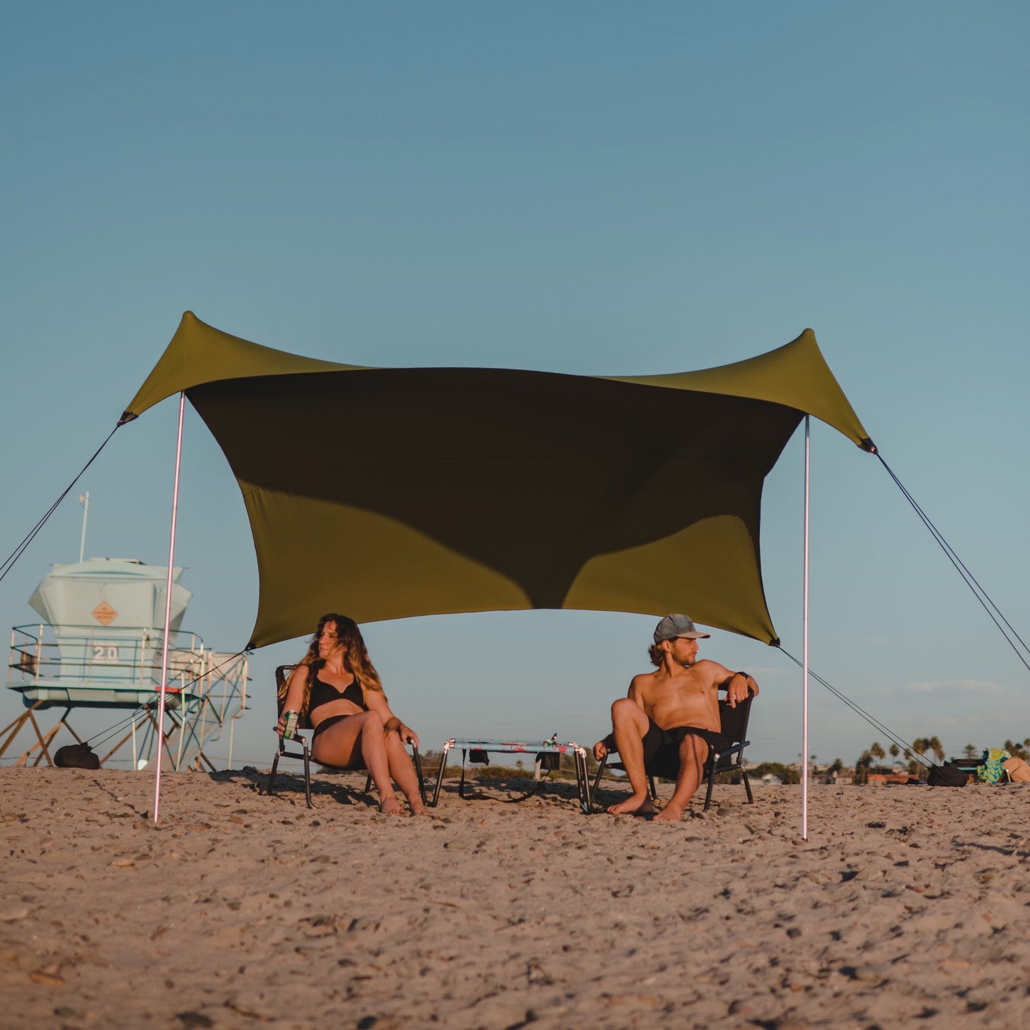 Two people sitting under a large green and black beach canopy on a sandy beach.