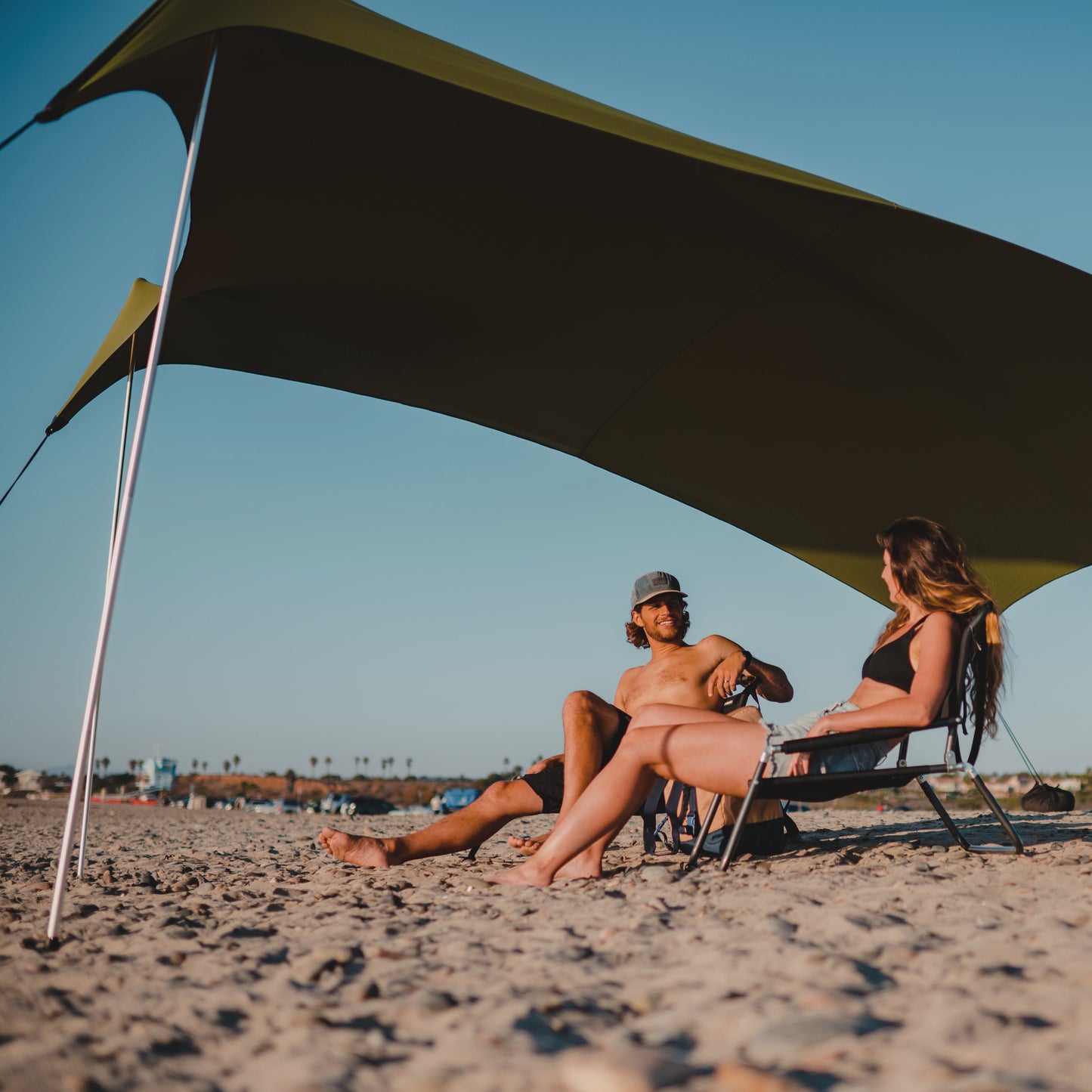 Two people sitting under a large green canopy on a sandy beach.