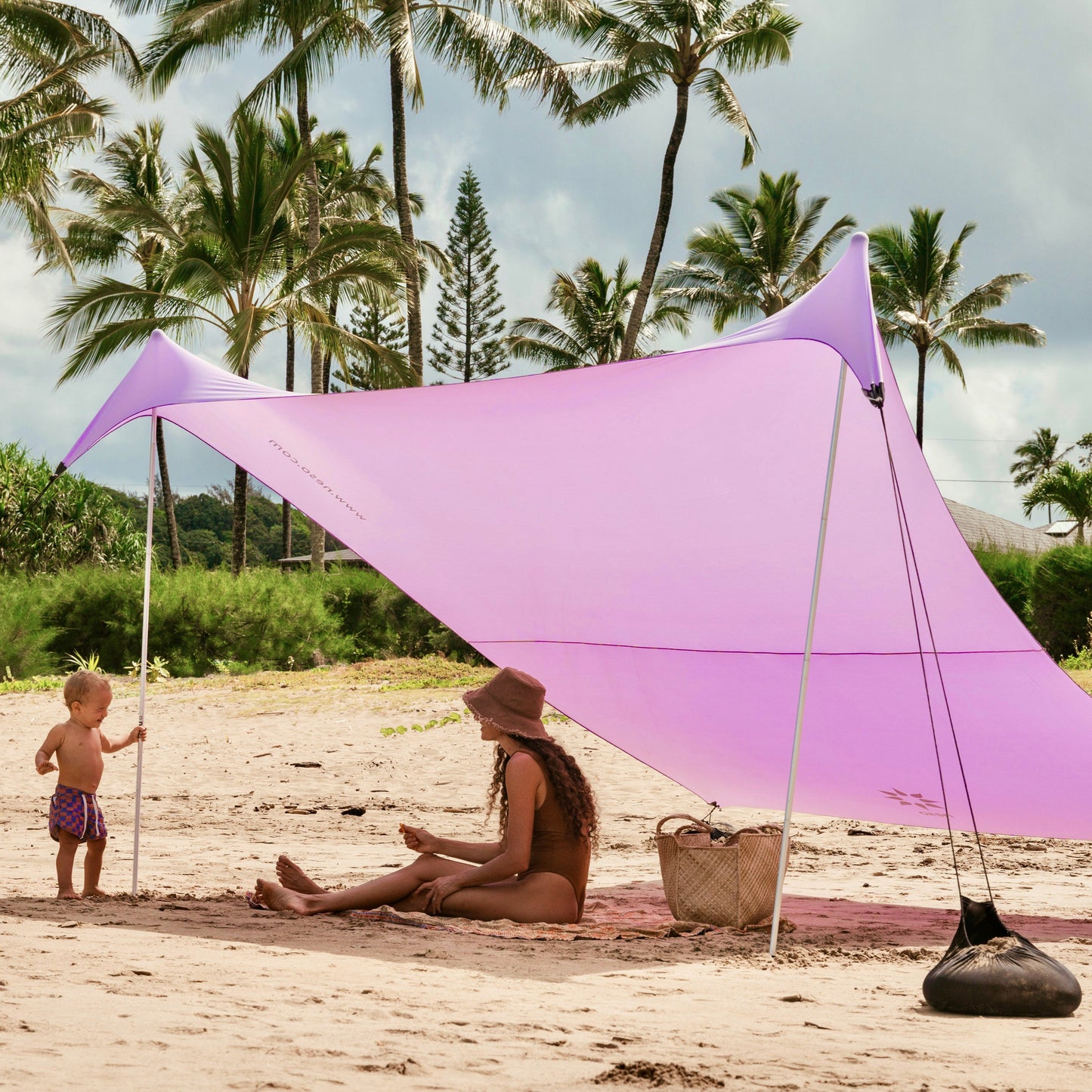 Woman and child under a purple NESO beach canopy on a sandy beach with palm trees in the background
