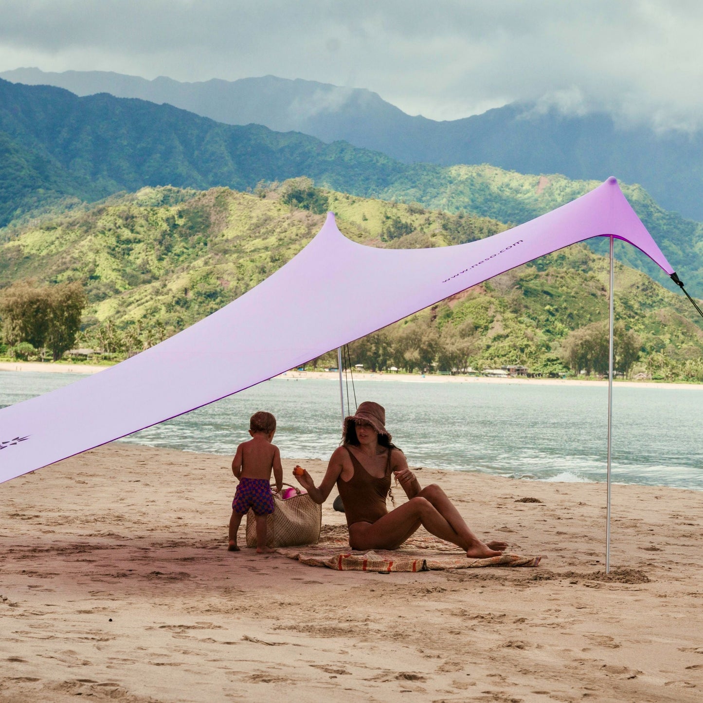 Woman and child sitting under a purple NESO beach canopy with water and mountains in the background.