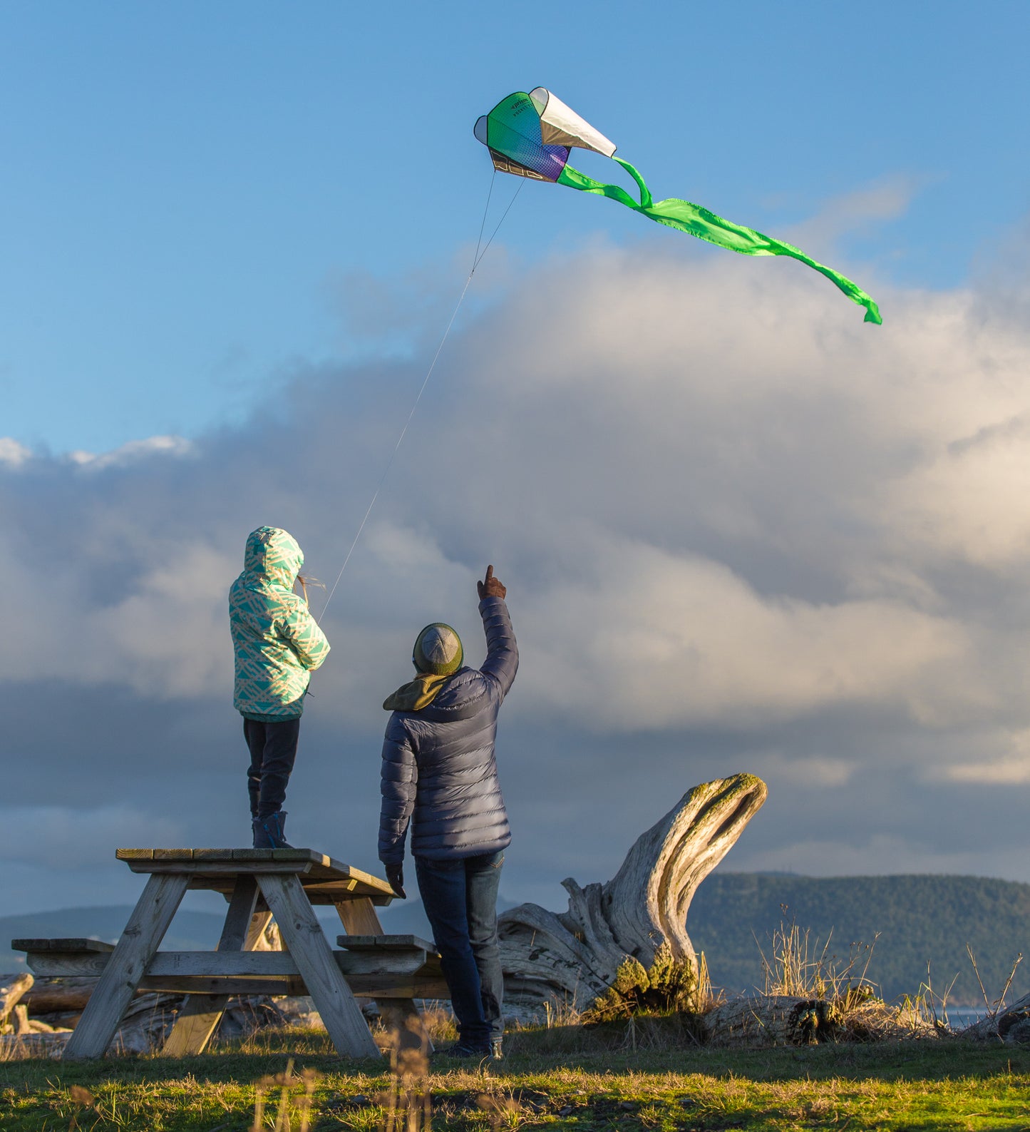 Two people flying a kite on a wooden bench with a scenic background