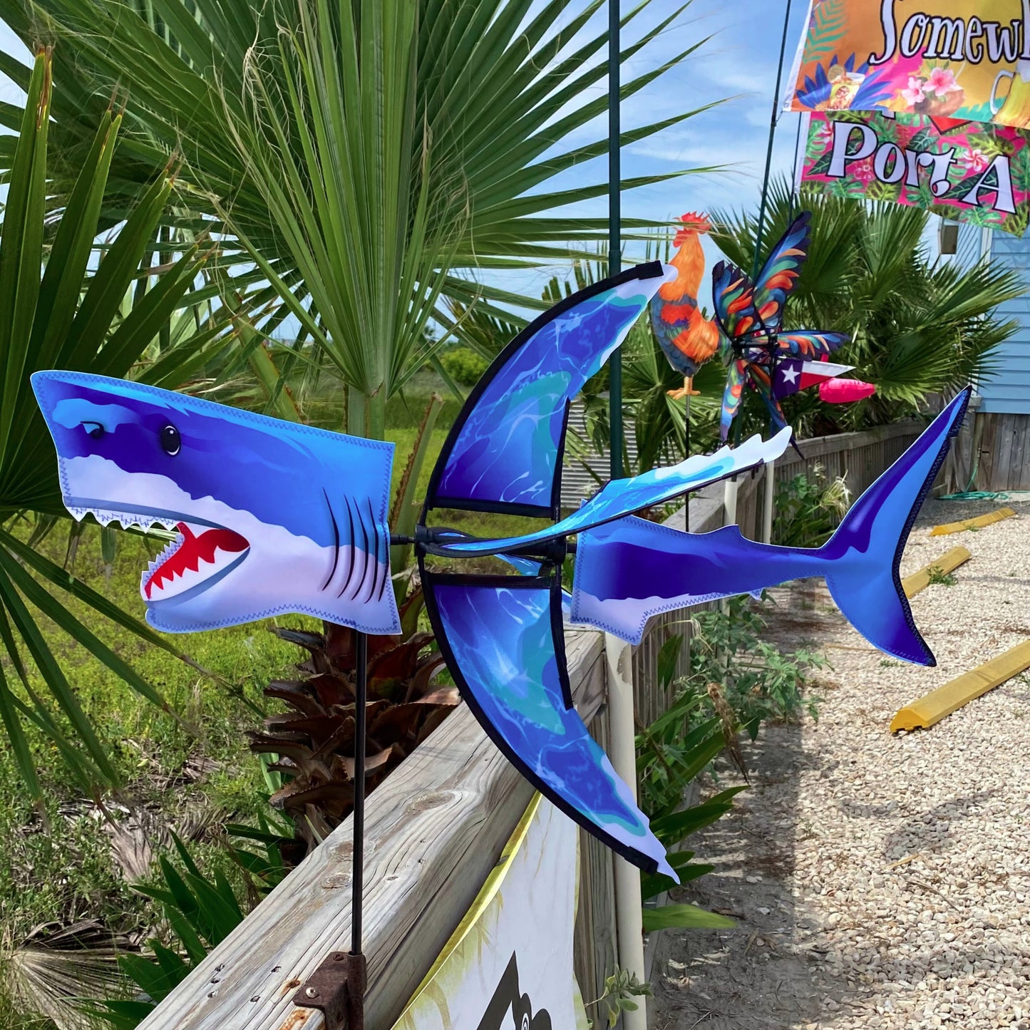 Blue shark-shaped wind spinner on a wooden post with palm trees and a colorful sign in the background.