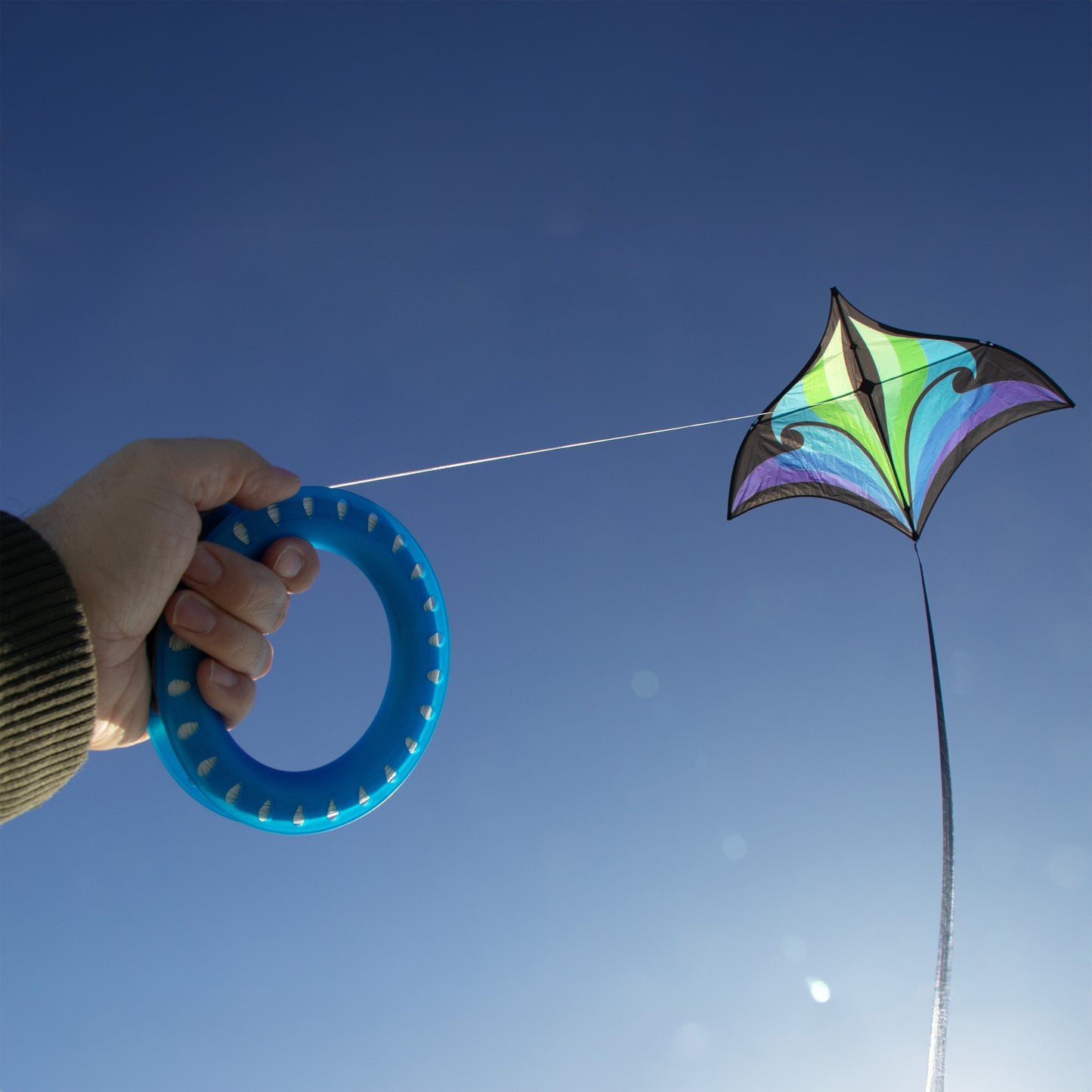 Hand holding a blue spool of string with a colorful kite flying against a clear blue sky.