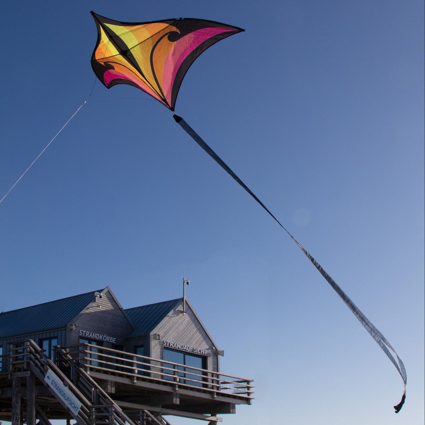 Colorful kite flying against a clear blue sky with a wooden structure in the background.