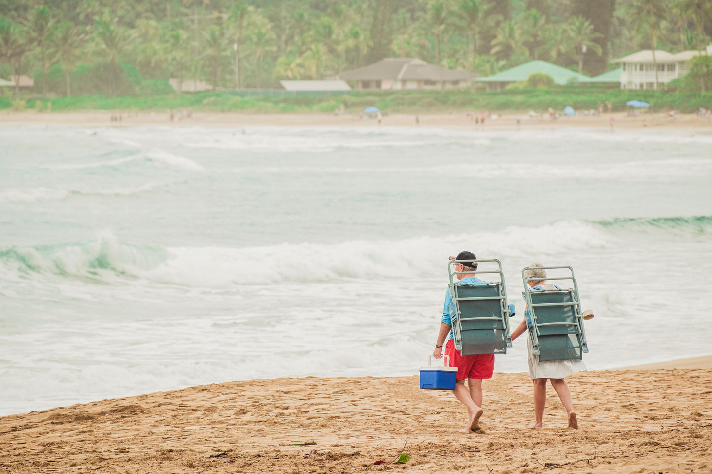 Two people walking on a sandy beach carrying large green beach chairs, with ocean waves and palm trees in the background.