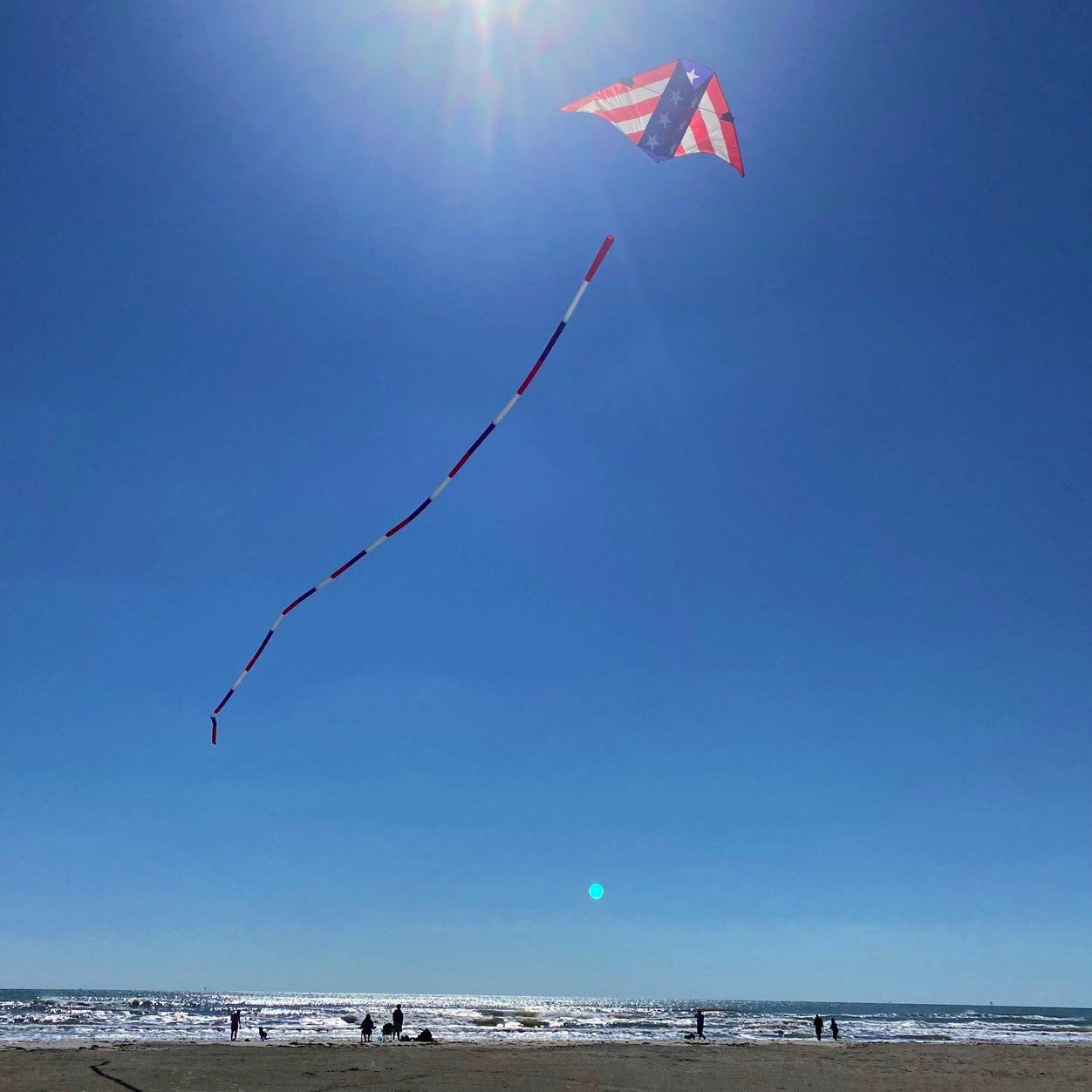 Kite flying on a beach with a clear blue sky and sun.