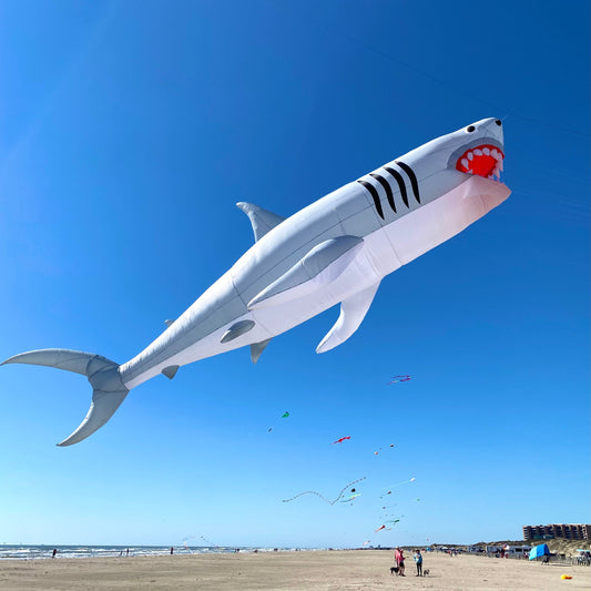 Shark-shaped kite flying high in the sky with a beach and people below.