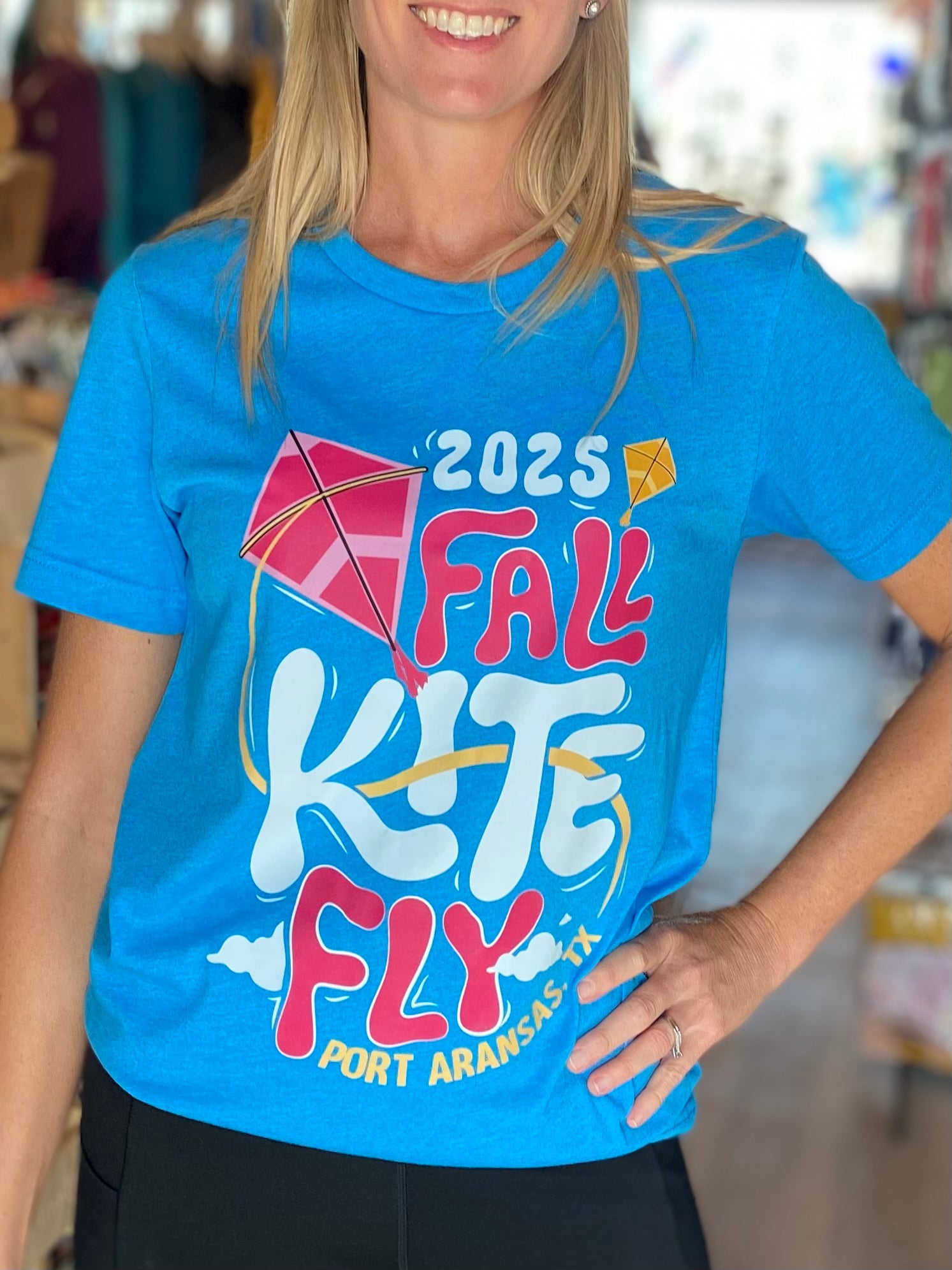 Person wearing a blue t-shirt with '2025 Fall Kite Fly Port Aransas, TX' text and graphics.