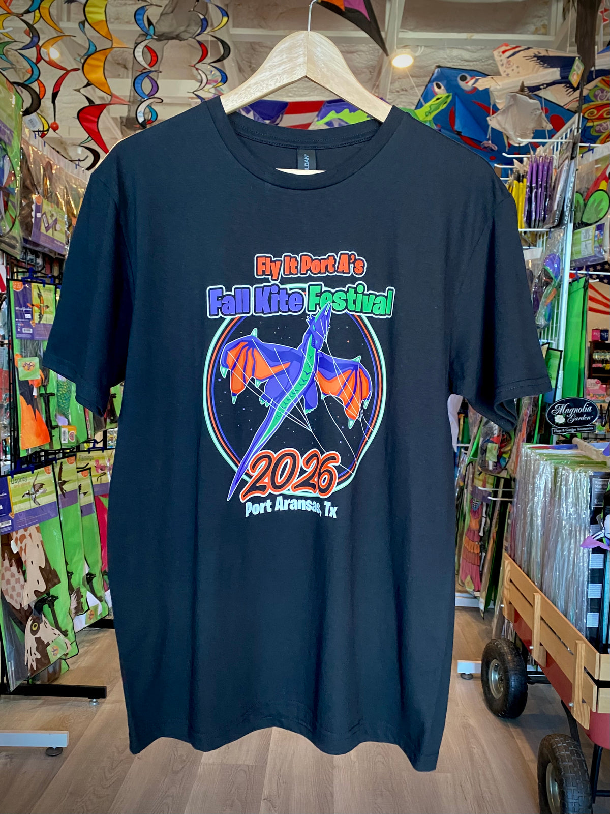 A black t-shirt hanging on a wooden hanger inside a retail store. The shirt features a colorful graphic print of a dragon with the text 'By It Port It's Fall Lake Festival 2026 Port Aransas, Tx'.