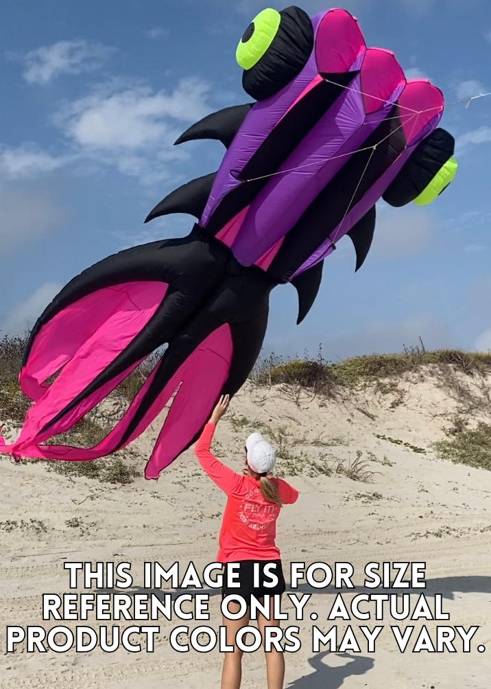 Person launching a large inflatable goldfish kite on a sandy beach with a blue sky.