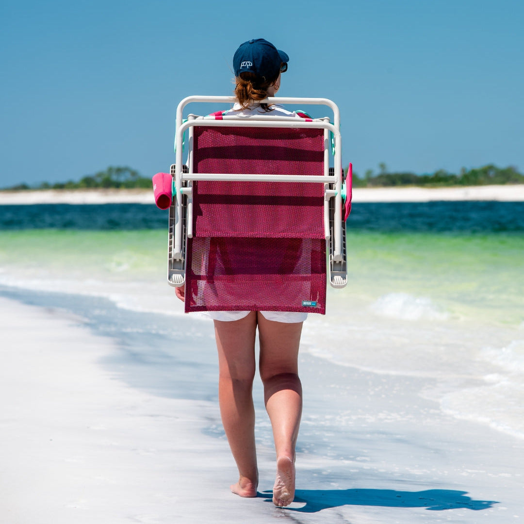 Person walking on a beach carrying a berry colored chair like a backpack. 