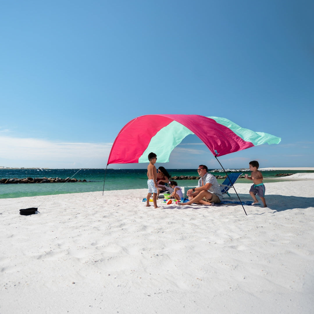 Family under a mint and berry colored shade on a sandy beach with ocean view