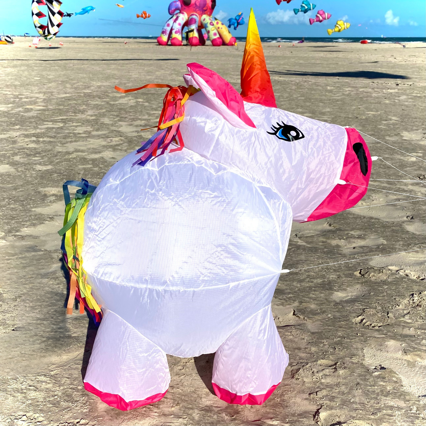 Colorful unicorn kite on a sandy beach with other kites in the background.