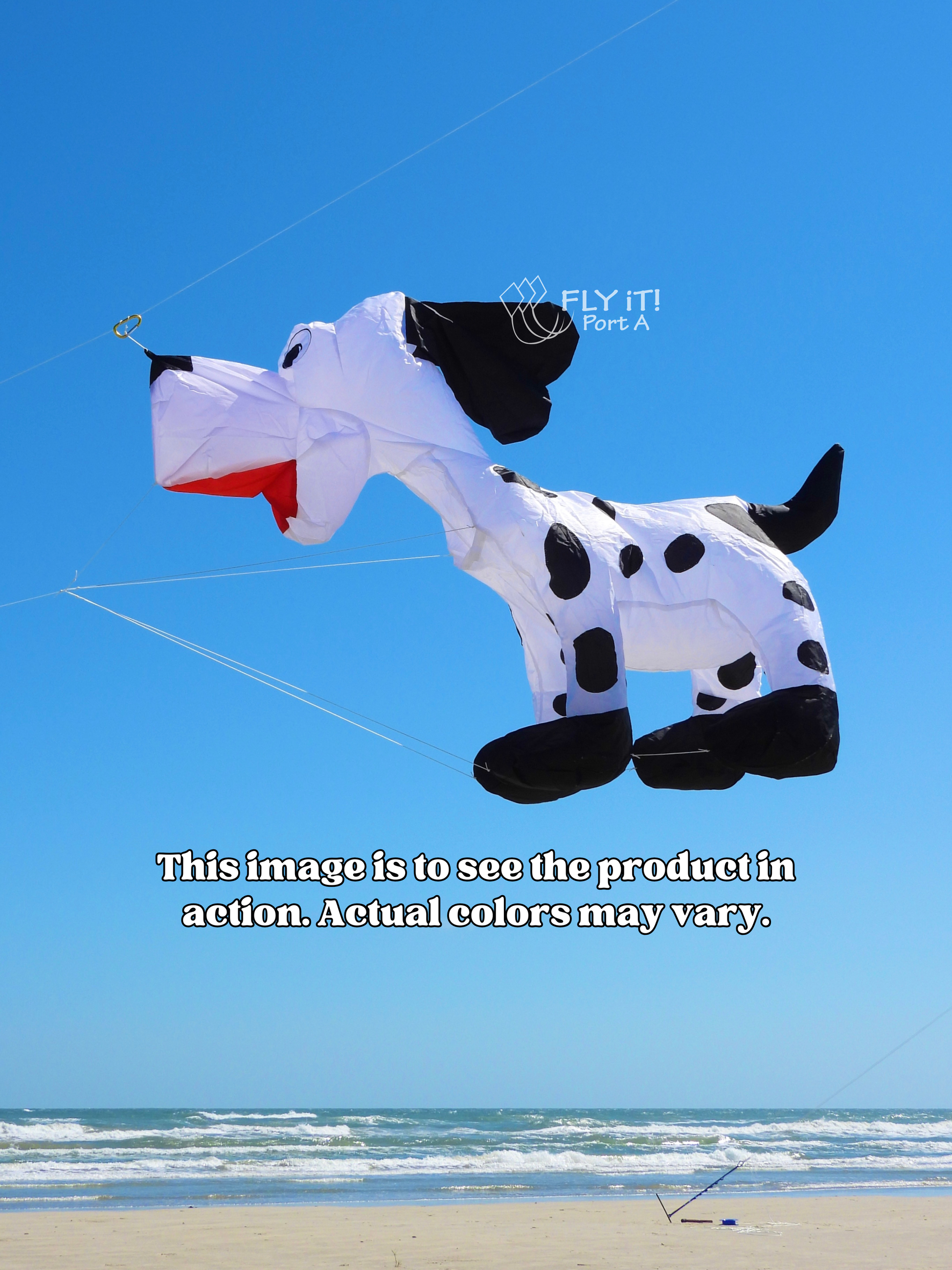 A large dog-shaped kite flying in the sky, designed like a Dalmatian with black spots on a white body and a red mouth. The background shows a bright blue sky and the ocean waves on the horizon, with a sandy beach at the bottom.