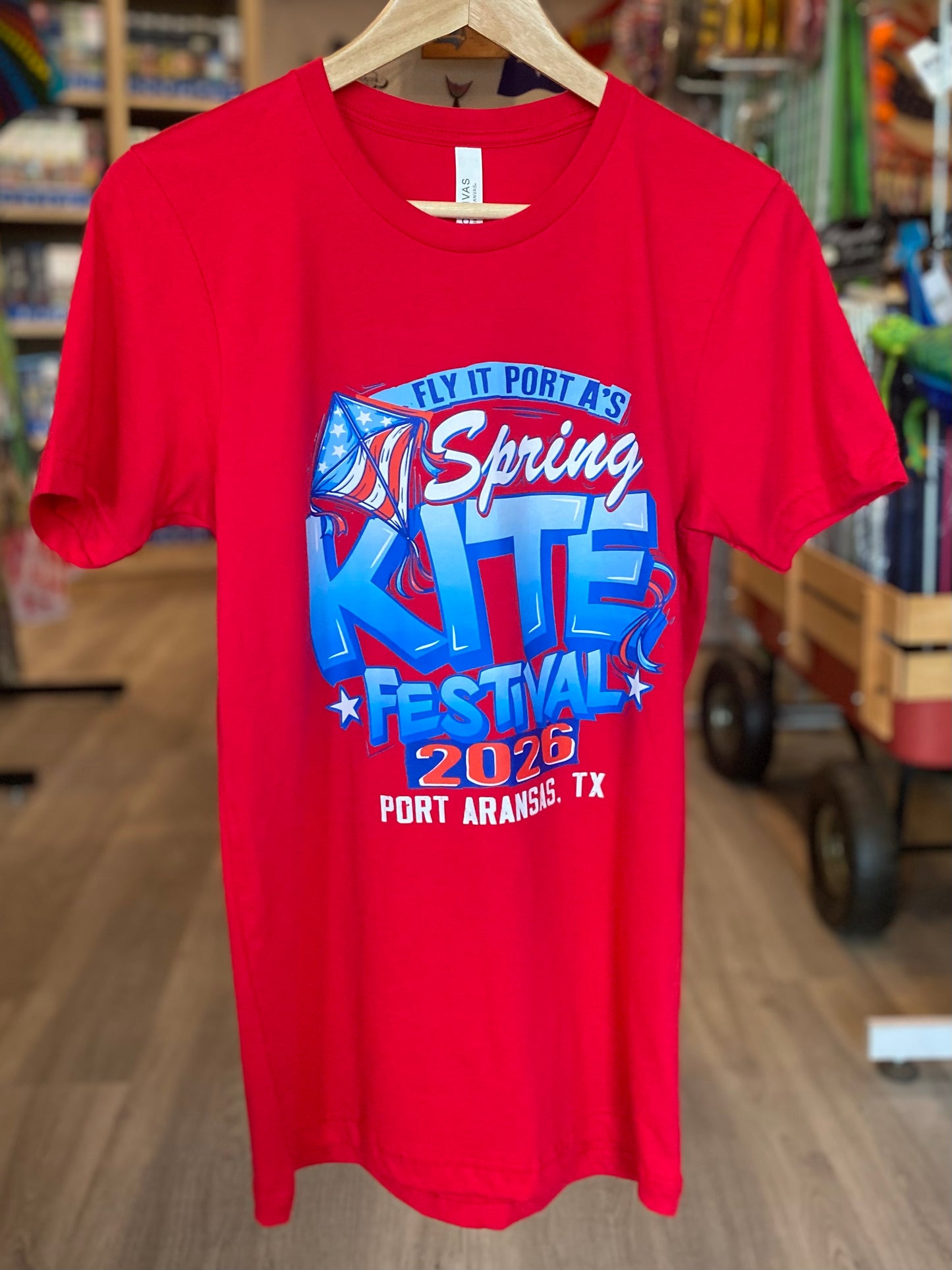 Red t-shirt with 'Spring Kite Festival 2026' design on a hanger in a store setting.
