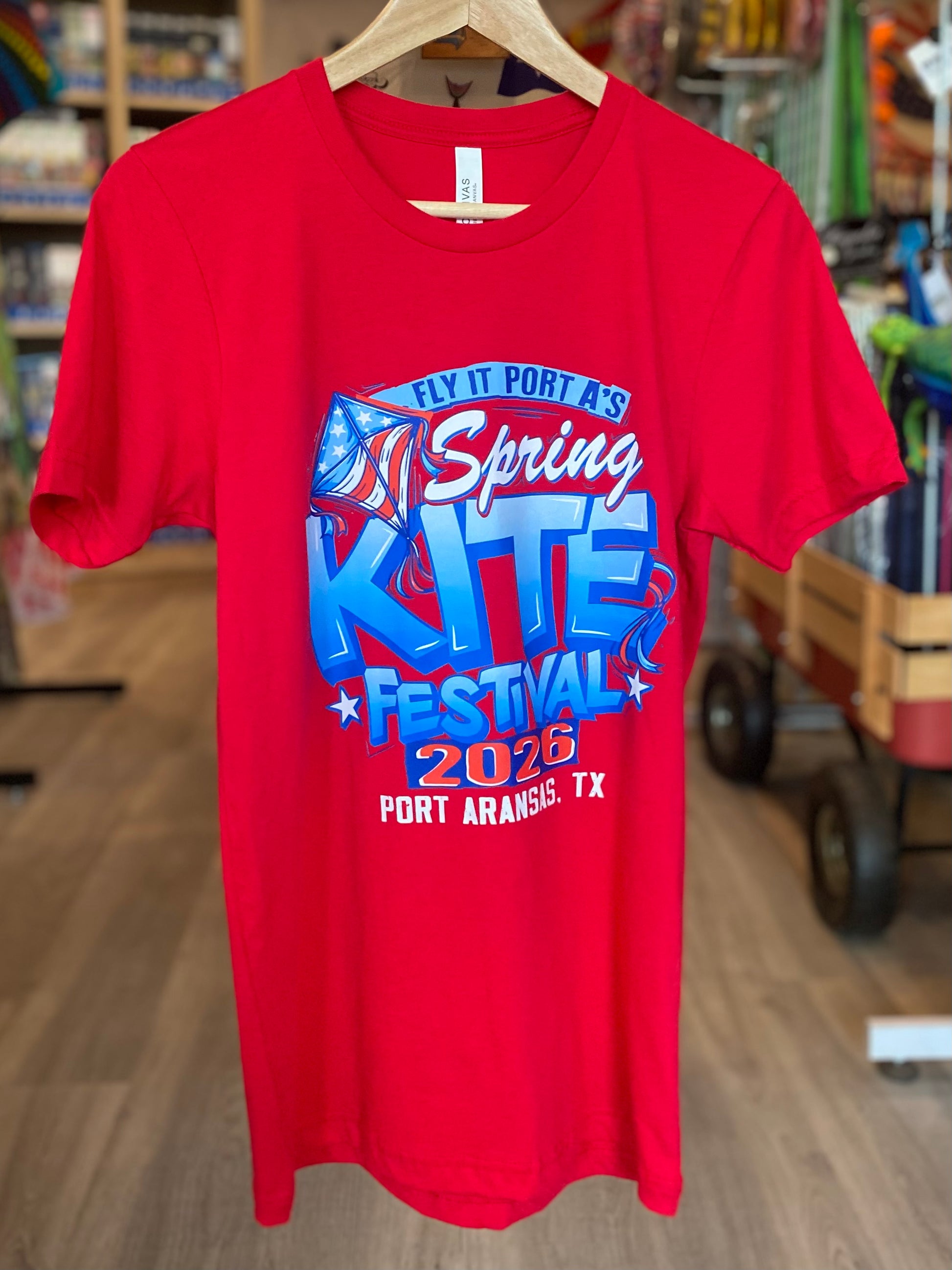 Red t-shirt with 'Spring Kite Festival 2026' design on a hanger in a store setting.