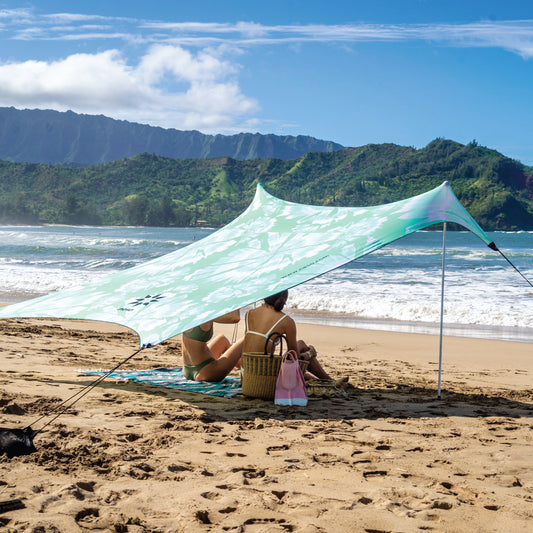Person sitting under a green and white NESO canopy on a sandy beach with ocean and mountains in the background