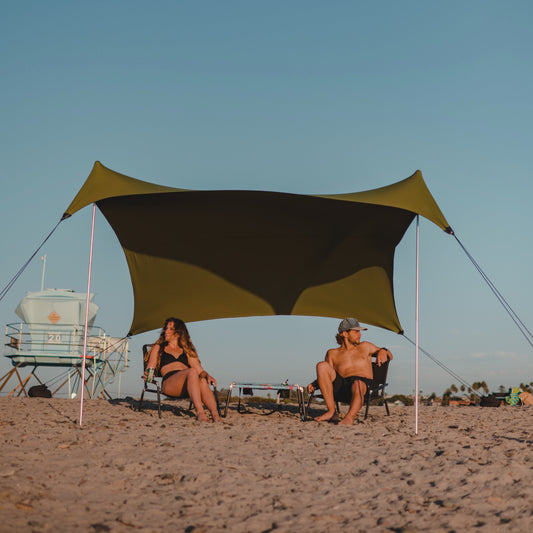 Two people sitting under a large green and black beach canopy on a sandy beach.