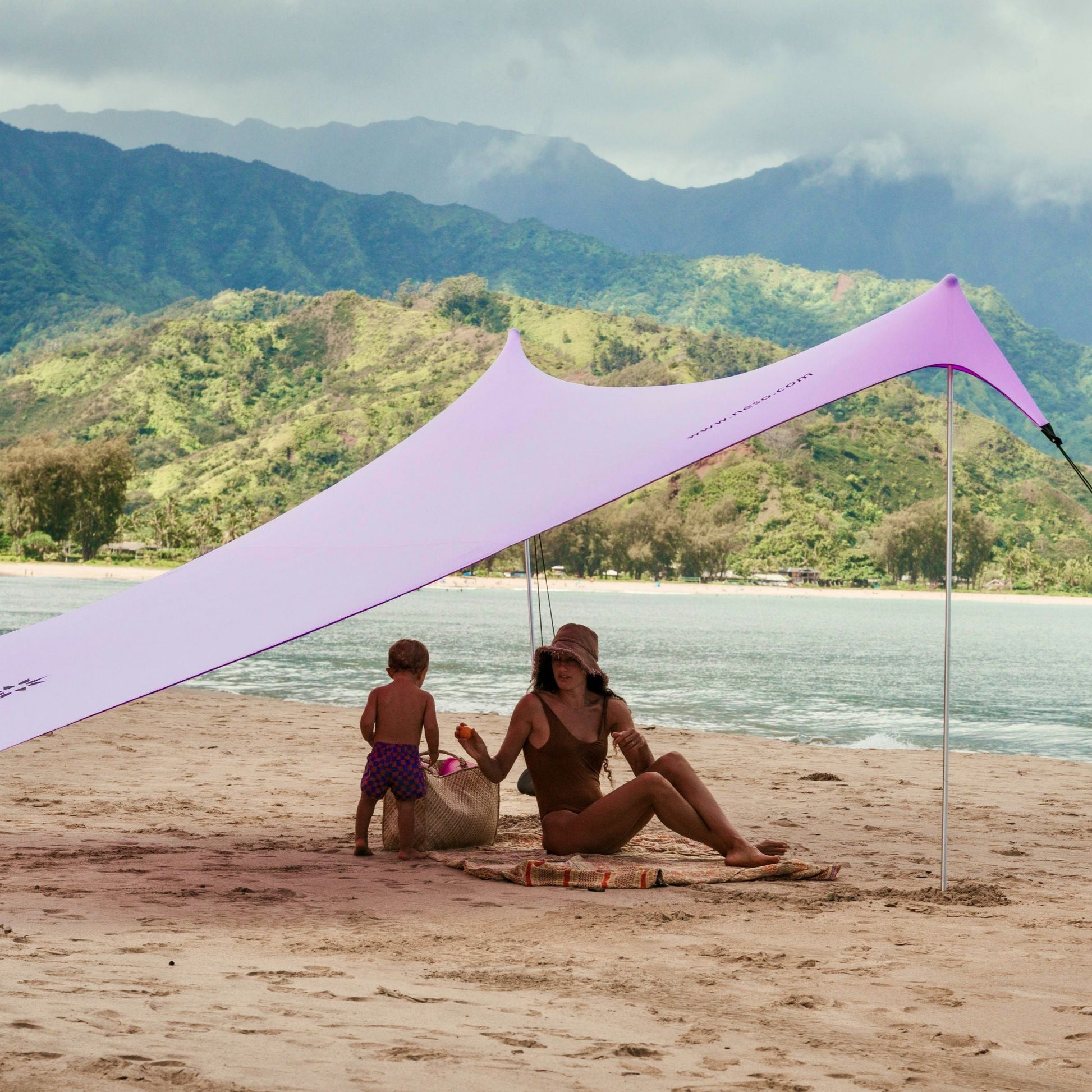 Woman and child sitting under a purple NESO beach canopy with water and mountains in the background.