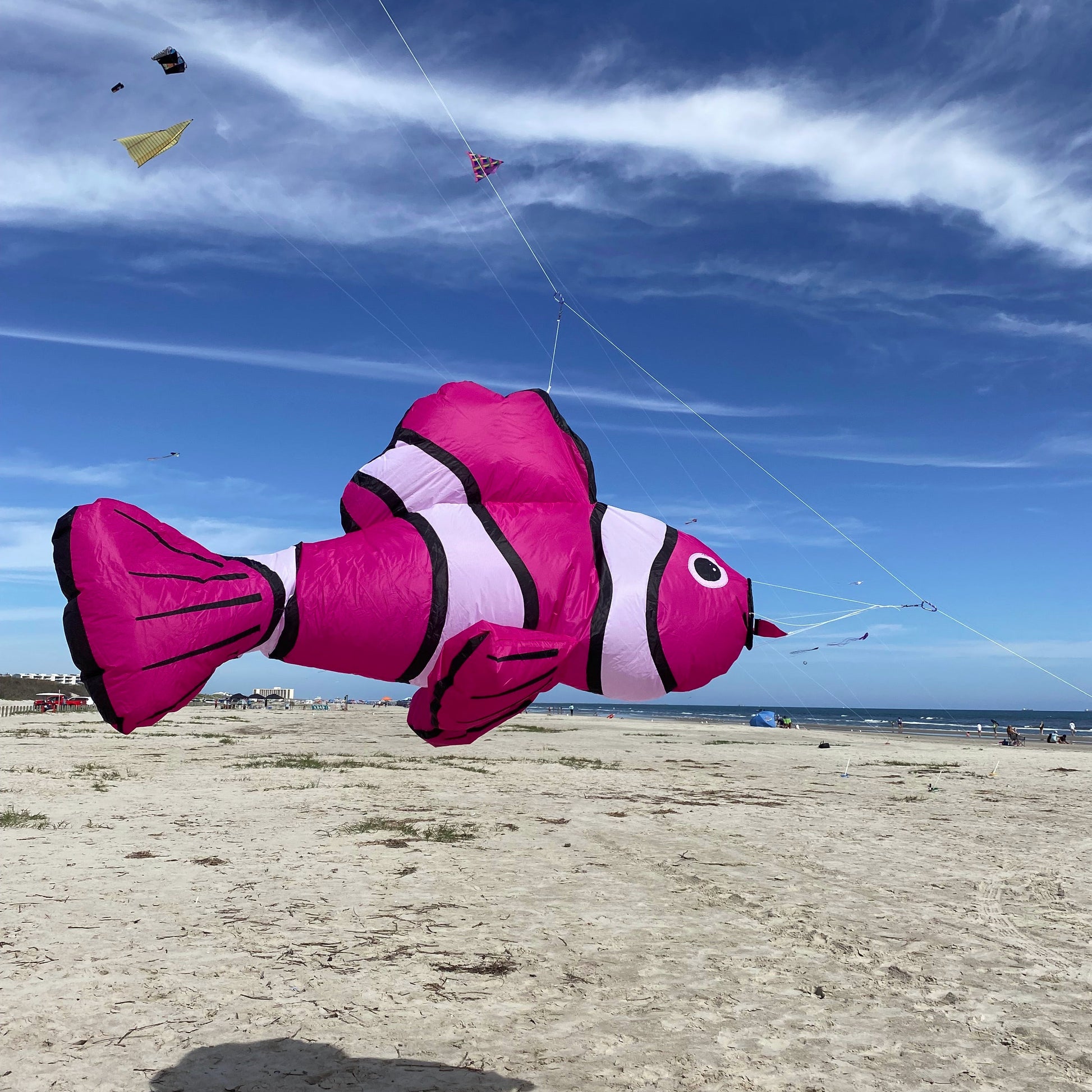 Pink fish-shaped kite flying against a blue sky with other kites on a sandy beach.