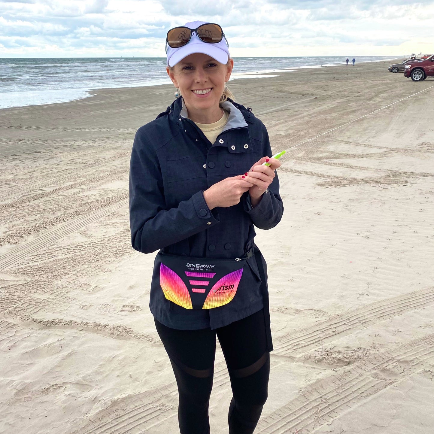 Person standing on a beach holding a small object, wearing a dark jacket with colorful kite bag around her waist.