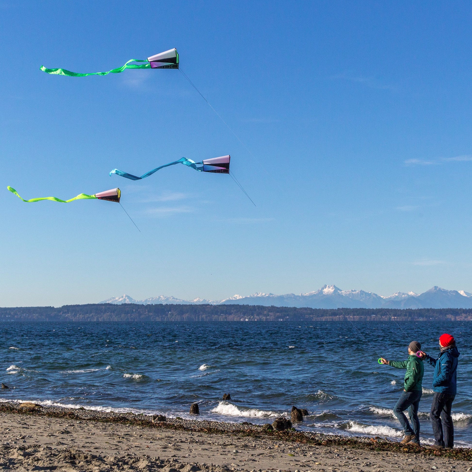 Two people flying kites on a beach with mountains in the background