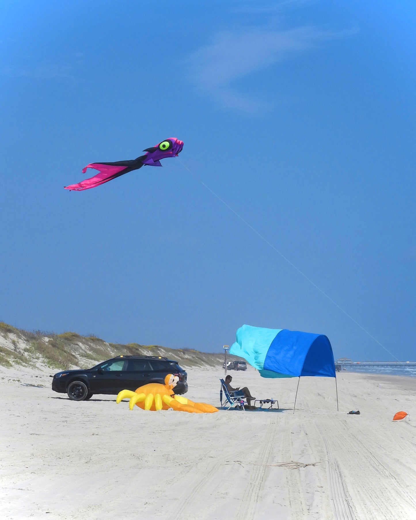 Beach scene with a large kite, car, and beach umbrella under a clear blue sky.