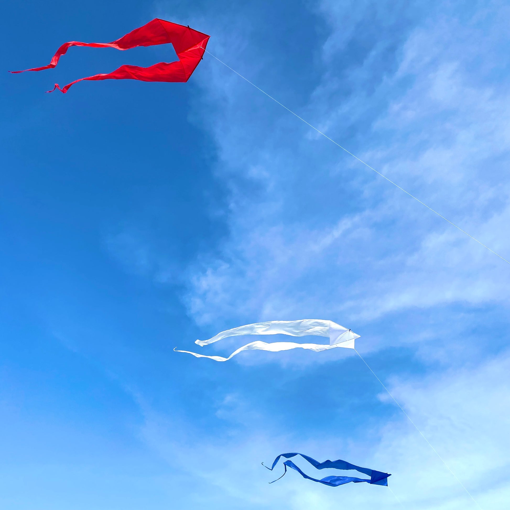 Red, white, and blue kites flying against a blue sky with some clouds.