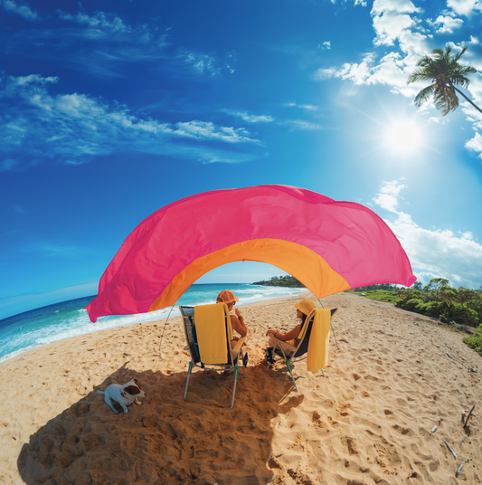 Two people sitting under a pink and orange beach canopy on a sandy beach with a clear blue sky.