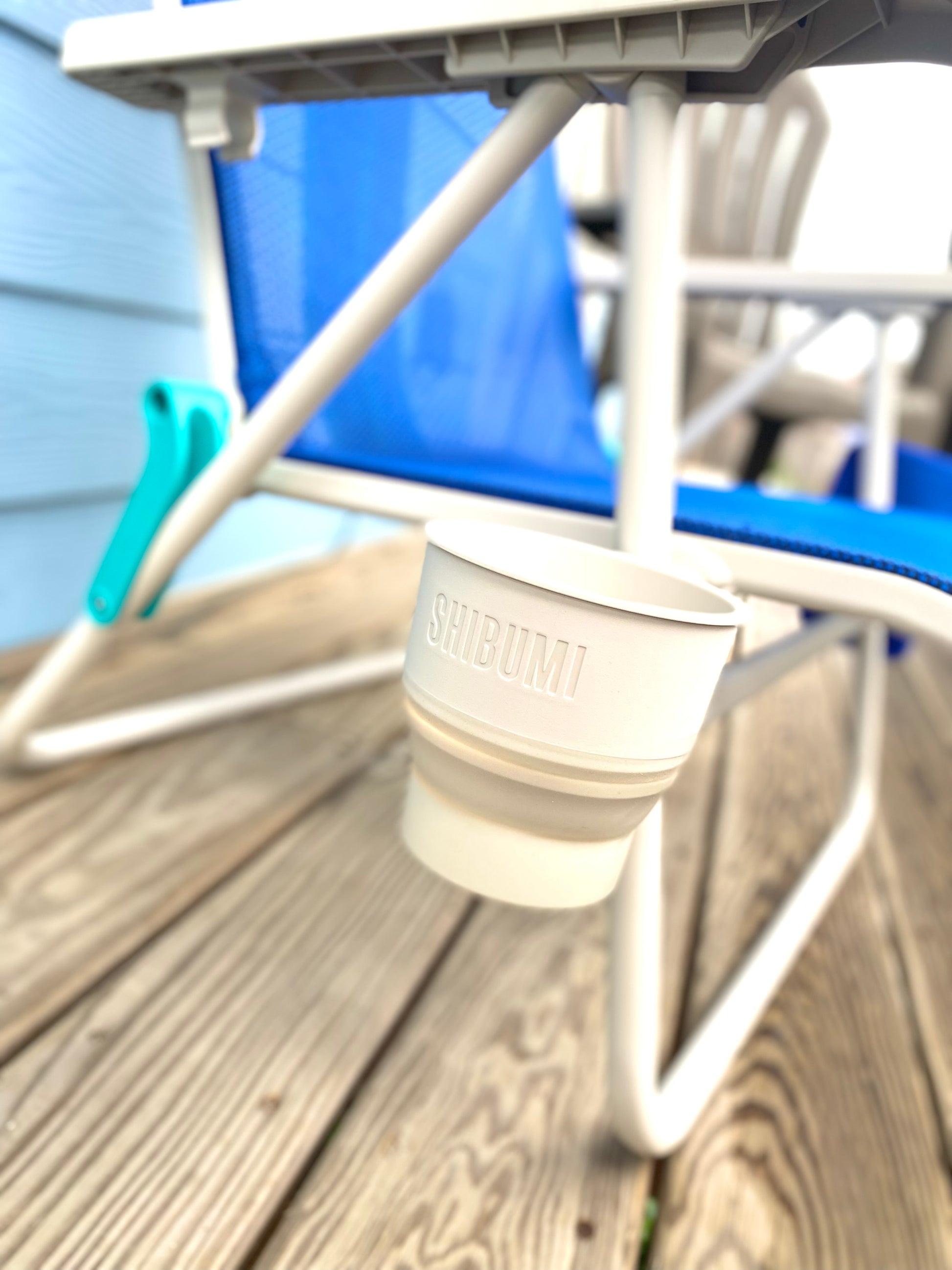 Beige cup with 'Shibumi' branding on a wooden deck with a blue chair in the background.