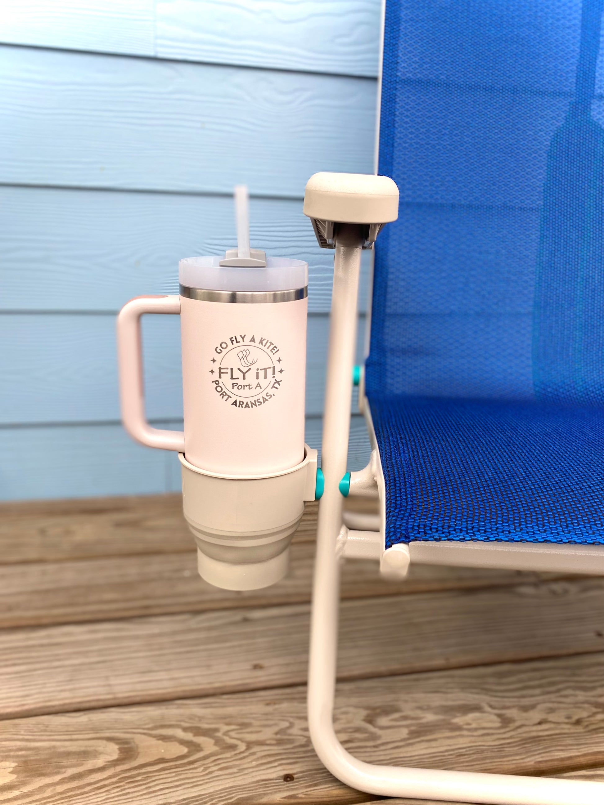 White tumbler with pink handle in a cup holder attached to a blue lounge chair on a wooden deck.