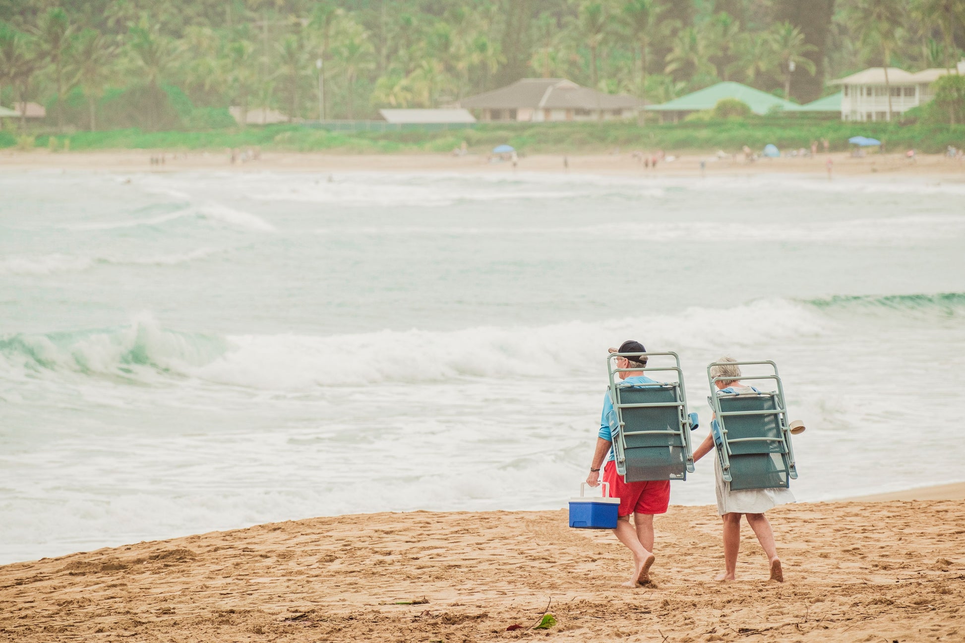 Two people walking on a sandy beach carrying large green beach chairs, with ocean waves and palm trees in the background.