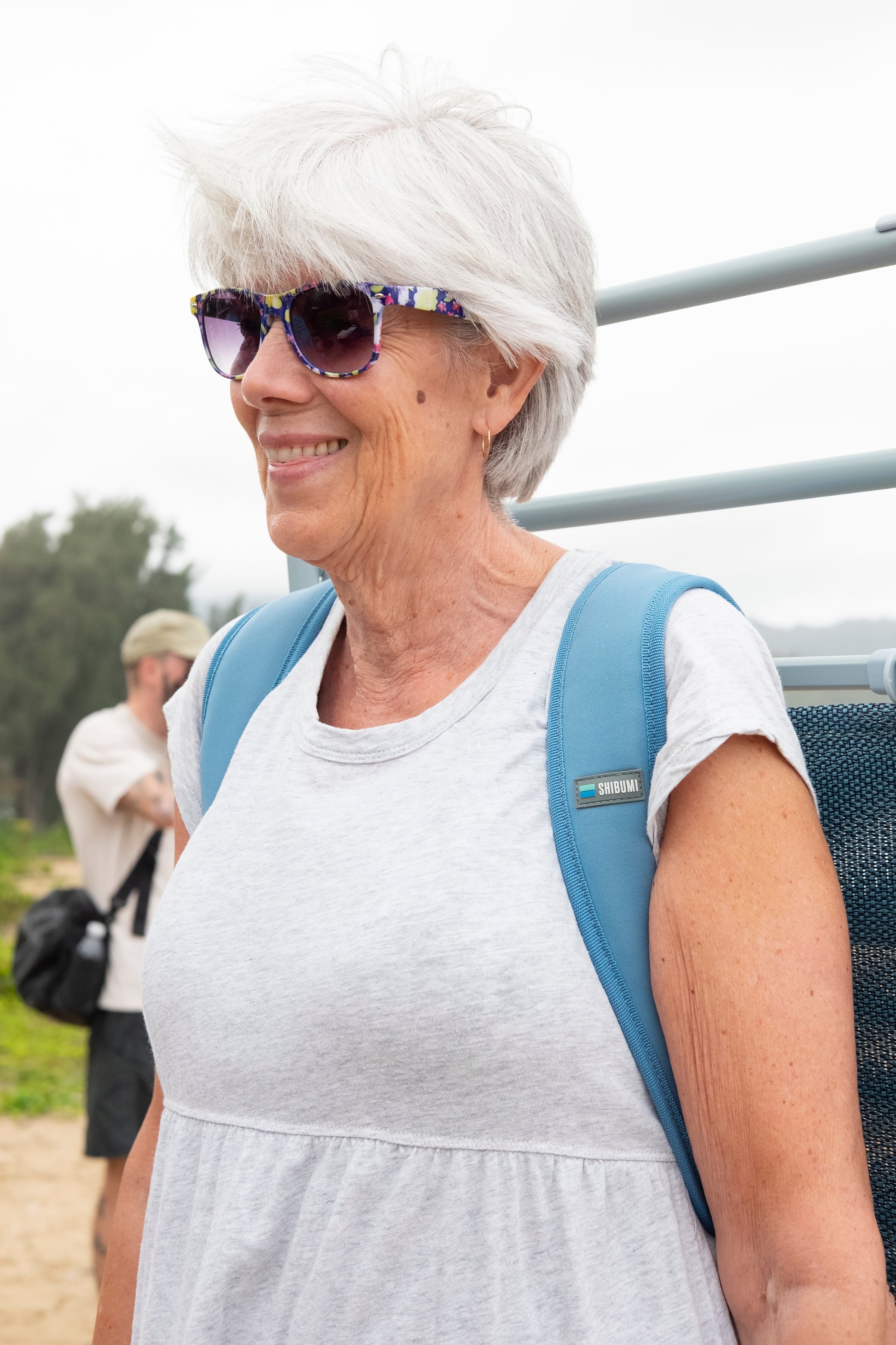 A smiling woman with short white hair and patterned sunglasses wears a light gray sleeveless top and a blue backpack outdoors.