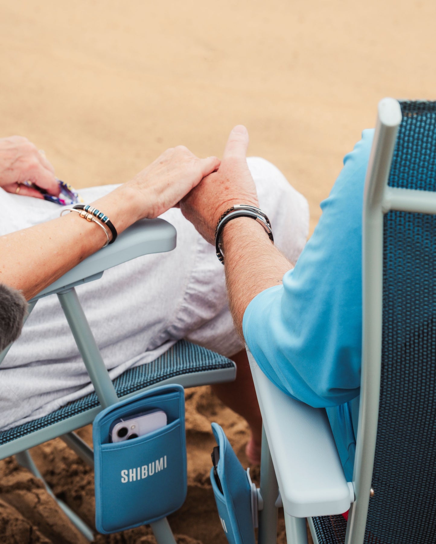 Close-up beach scene showing two people sitting in blue mesh folding chairs on sand, with one chair side pocket holding a white phone and the word SHIBUMI printed on it.