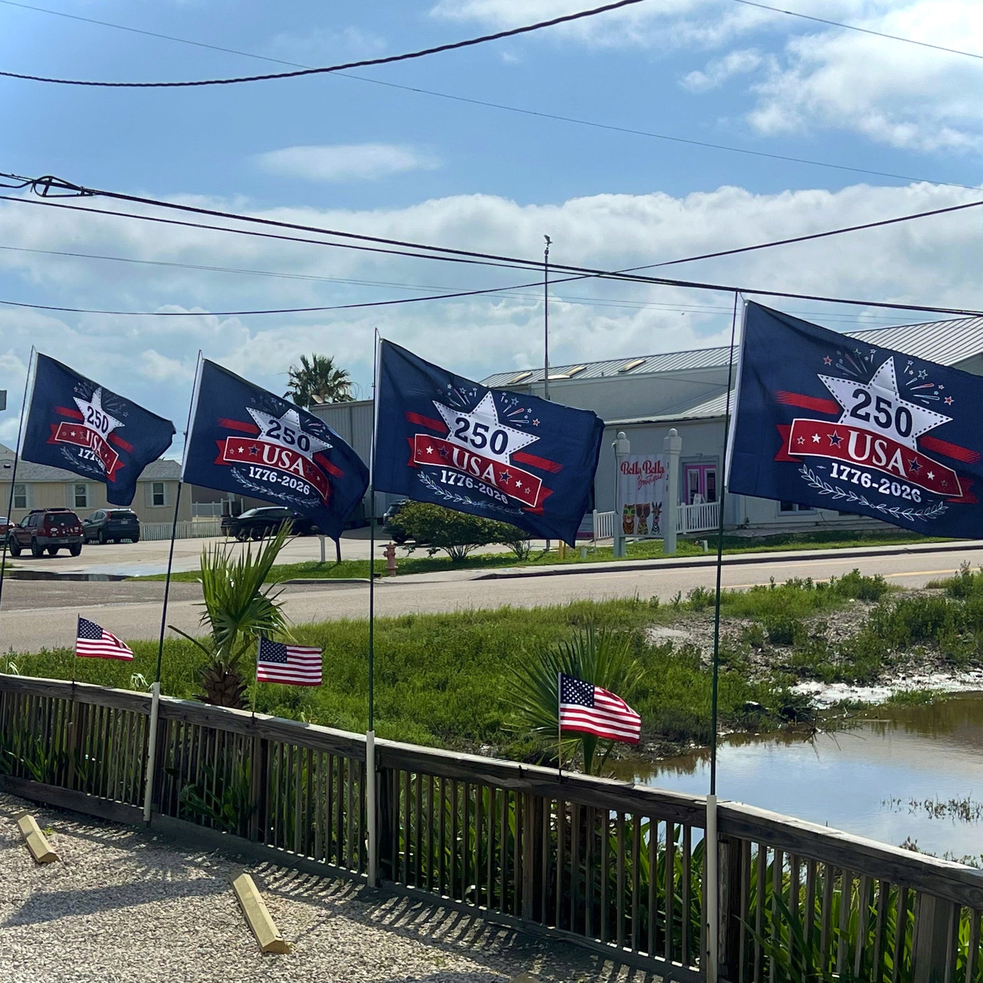 Flags with 'USA' and '250' text on a fence by a road with buildings in the background.