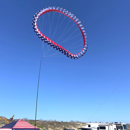 Red, white, and blue ring-like kite flying high in a clear blue sky with people and vehicles on the ground.