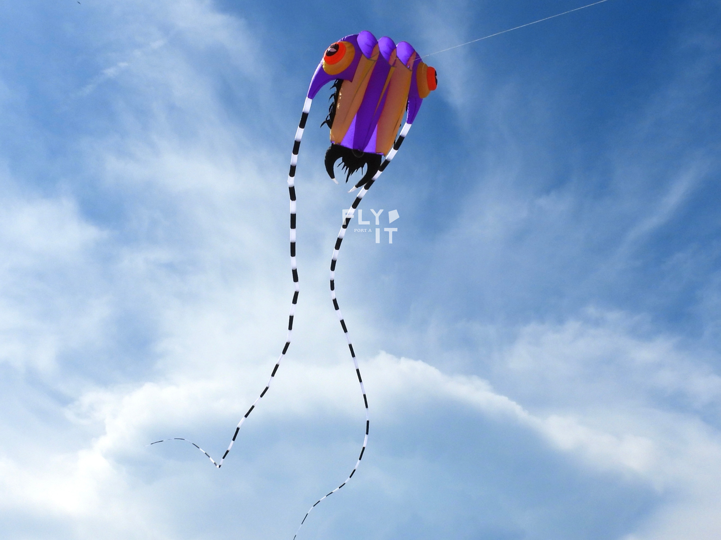 A colorful trilobite kite with purple, orange, and black colors flying in a blue sky with light clouds. The kite has long black-and-white striped tails and a small white 'FLY IT' watermark near the center.