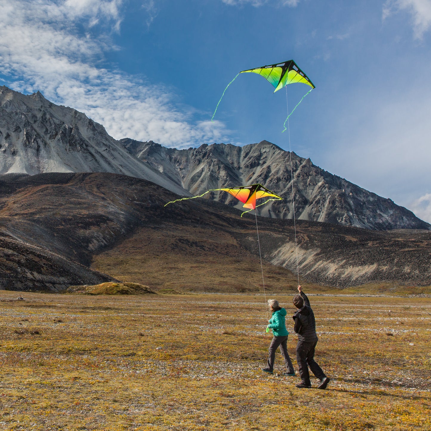 Two people flying kites in a mountainous landscape with a clear blue sky.