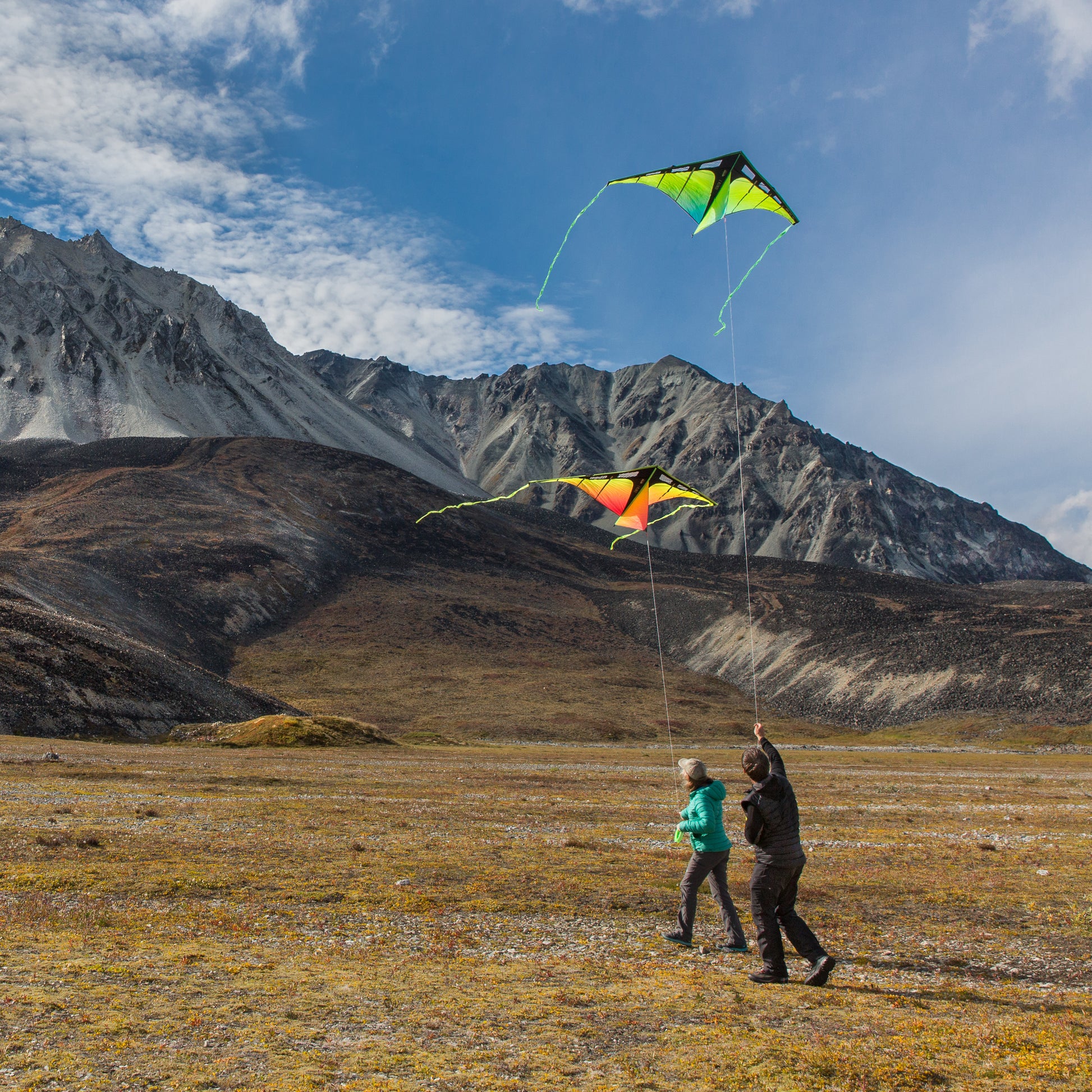 Two people flying kites in a mountainous landscape with a clear blue sky.